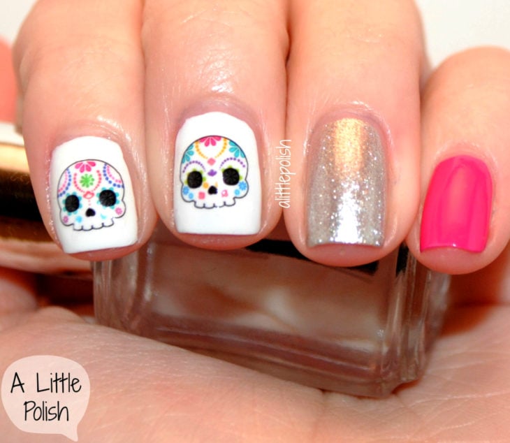 Manicura de Día de Muertos; uñas pintadas diferente, con esmalte blanco, plateado con glitter y rosa, con diseño de calavera de dulce con flores