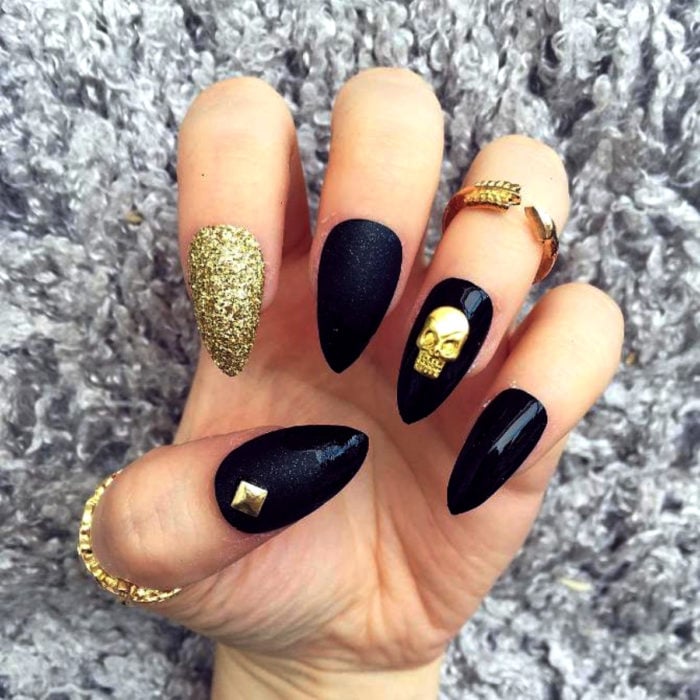 Manicura de Día de Muertos; uñas largas en forma de almendra, pintadas diferente con esmalte negro y dorado con glitter, y con adorno de cráneo y un estoperol