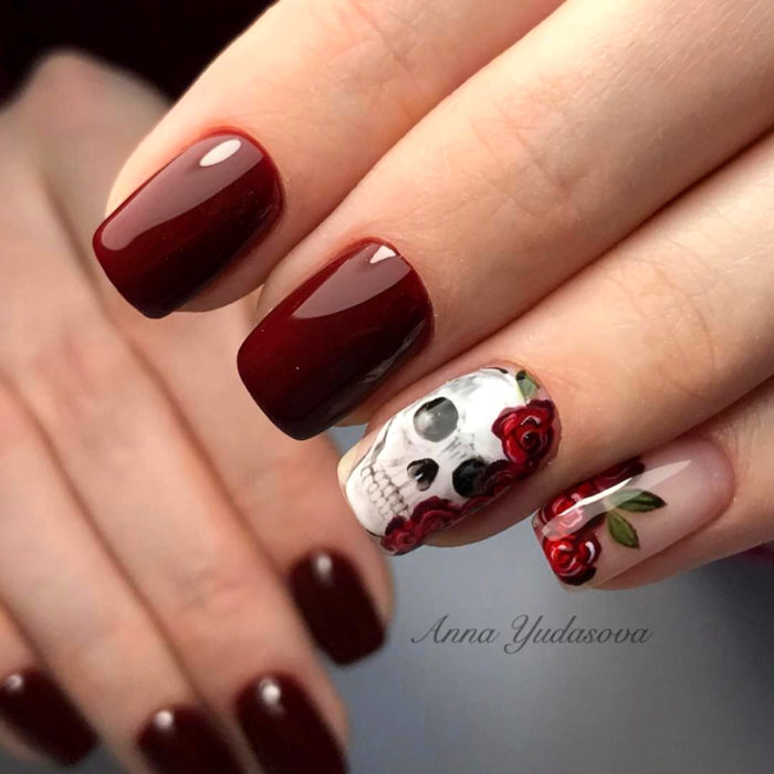 Manicura de Día de Muertos; uñas largas cuadradas, pintadas con esmalte color rojo vino y diseño de cráneo con flores