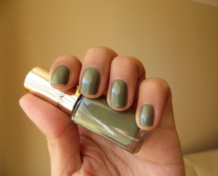 Chica sosteniendo un esmalte de color verde olivo mientras enseña sus uñas del mismo tono 