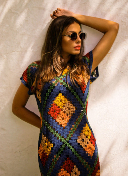 Mujer con lentes de sol, recargada en la pared, con vestido tejido de colores, azul, verde, amarillo, rojo, rosa y blanco