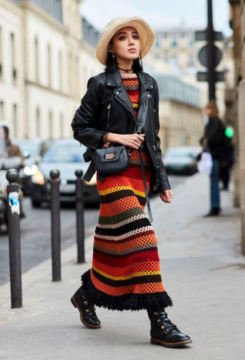 Mujer caminando en la calle, con cabello negro a los hombros, sombrero, maxivestido tejido de colores rojo, anaranjado, amarillo, blando, verde y negro con chaqueta de cuero y botas de trabajo