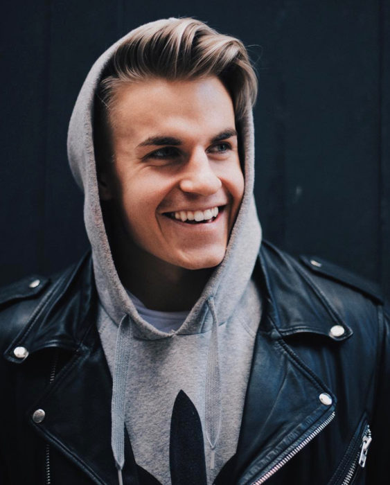 Finlandia te paga por tener bebés; guapo chico finlandés sonriendo, hombre rubio con chamarra de cuero negro y sudadera gris