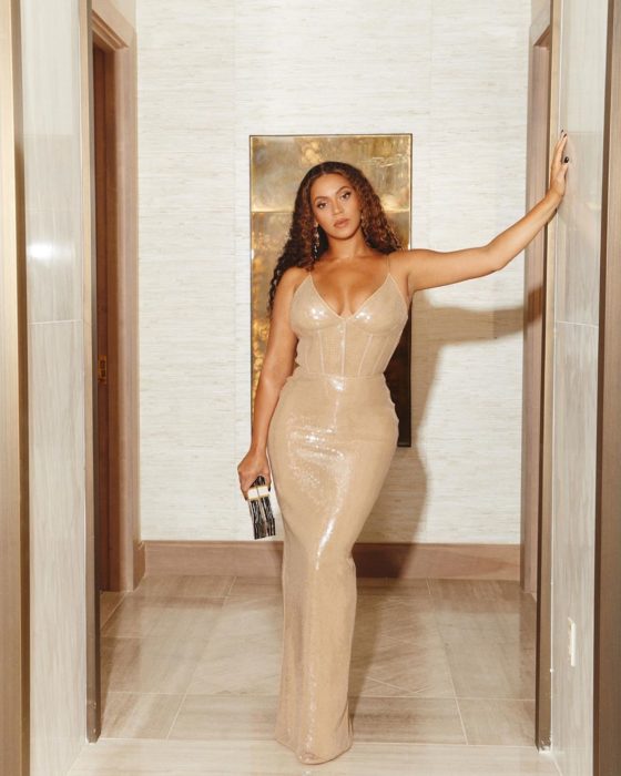 Beyoncé recargada en el marco de una puerta usando un vestido de color dorado con el cabello rizado