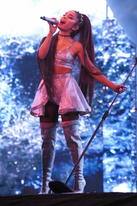 Ariana Grande en el escenario cantando mientras usa un vestido de color plateado y botas altas 