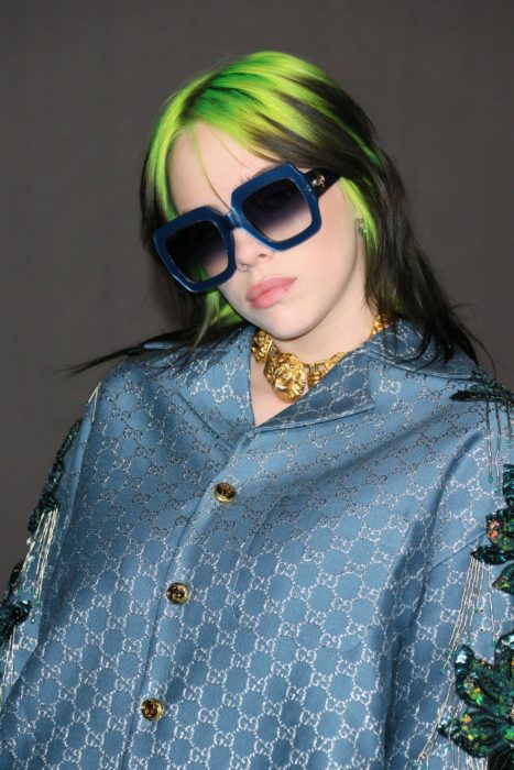 Cantante Billie Eilish usando un saco azul, lentes de sol azul y el cabello teñido de color verde 