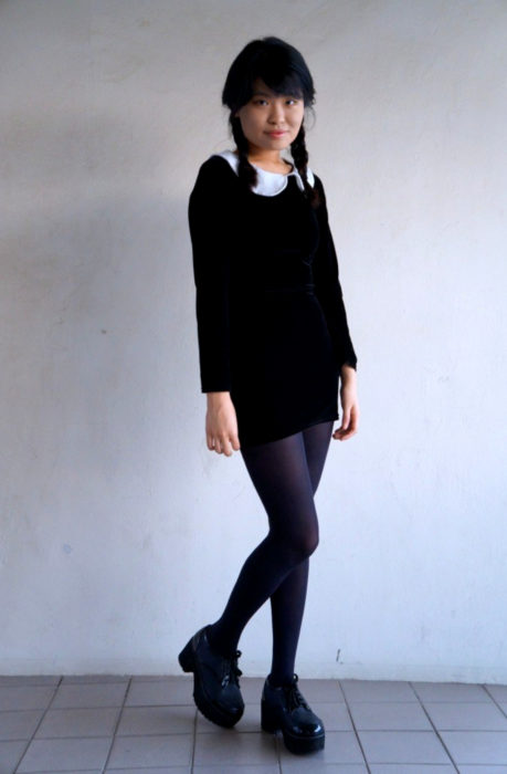 Chica asiática vestida como Merlina Adams; vestido negro, corto, con cuello blanco, zapatos de plataforma y peinado de trenzas