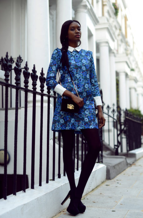 Chica morena vestida de Merlina Adams; vestido azul, de flores, con cuello blanco, medias negras y botas de tacón de aguja