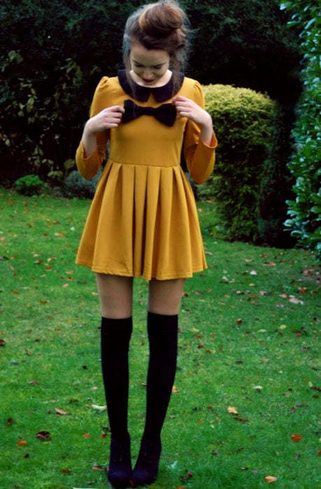 Chica vestida de Merlina Adams; vestido amarillo mostaza con moño negro, medias, botines, con peinado de chongo alto