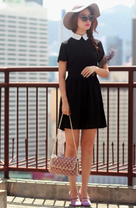 Chica vestida de Merlina Adams; vestido negro con cuello blanco, zapatos morados, sombrero lila, con lavanda en la mano y con lentes de sol