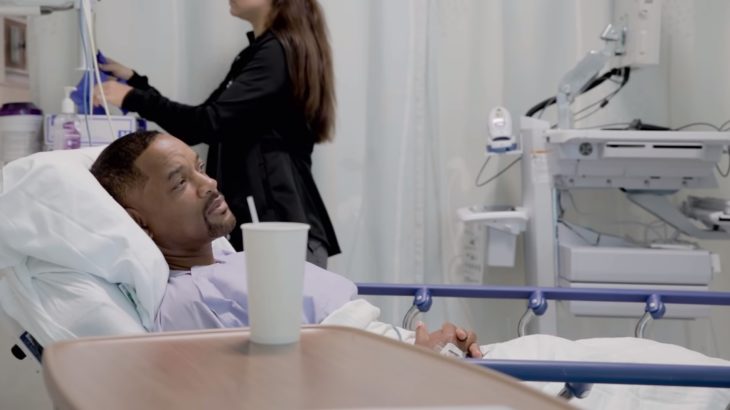 Will Smith en bata para un examen médico, recostado en una camilla