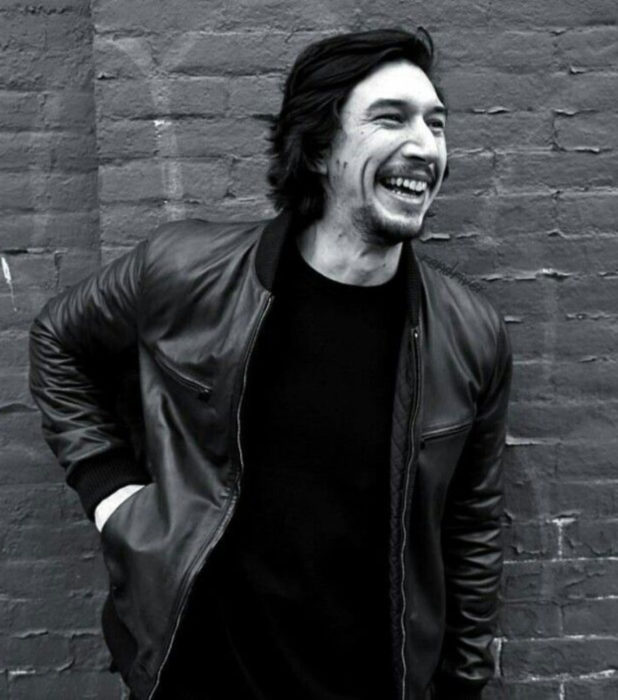 Actor Adam Driver; fotografía en blanco y negro de hombre recargado en la pared sonriendo, con chamarra de cuero, cabello largo y oscuro, barba y bigote