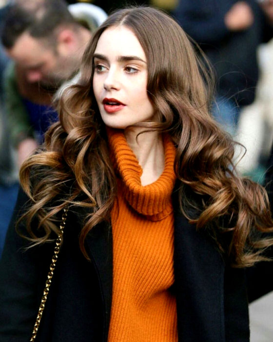 Peinados de Lily Collins para Año Nuevo; mujer de cabello castaño, largo, ondulado, con suéter de cuello de tortuga anaranjado