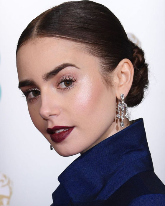 Peinados de Lily Collins para Año Nuevo; mujer de cabello lacio, castaño, recogido en un chongo bajo, maquillaje natural y labios color vino