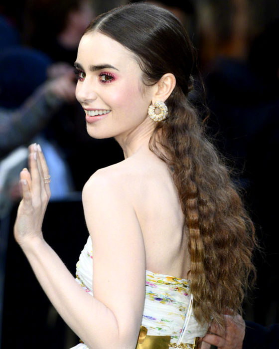 Peinados de Lily Collins para Año Nuevo; mujer de cabello largo, castaño, ondulado con wafflera, agarrado en media cola, con maquillaje natural y aretes grandes