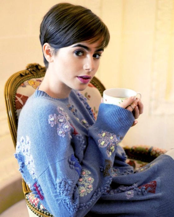 Peinados de Lily Collins para Año Nuevo; mujer de cabello castaño y lacio con corte pixie, con suéter holgado y tejido color azul, sentada en sillón vintage bebiendo una taza de té