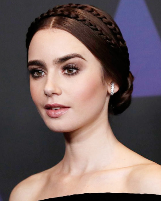 Peinados de Lily Collins para Año Nuevo; mujer de cabello castaño oscuro, con peinado de chongo y trenzas para fiesta, maquillaje negro natural