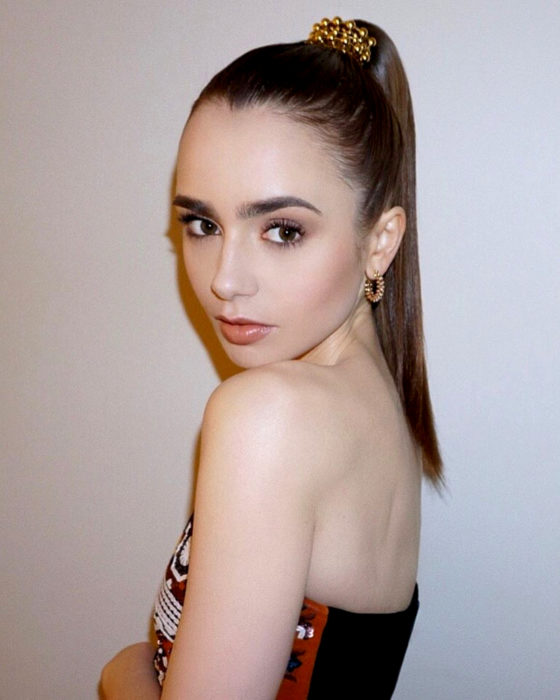 Peinados de Lily Collins para Año Nuevo; mujer de cabello largo, castaño y lacio, peinado con coleta alta con broche dorado, cejas prominentes