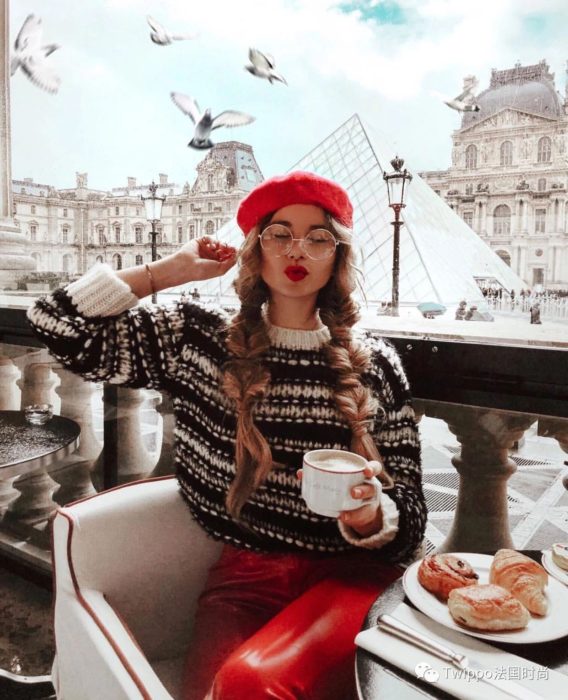 Chica sentada en un café de París usando un feo suéter navideño mientras toma un café 