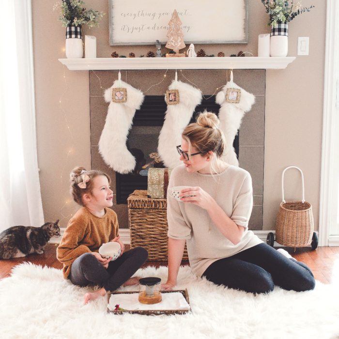 Mamá e hija sentadas en la alfombra, bebiendo te y platicando frente a la chimenea; botas colgadas y gato atigrado 