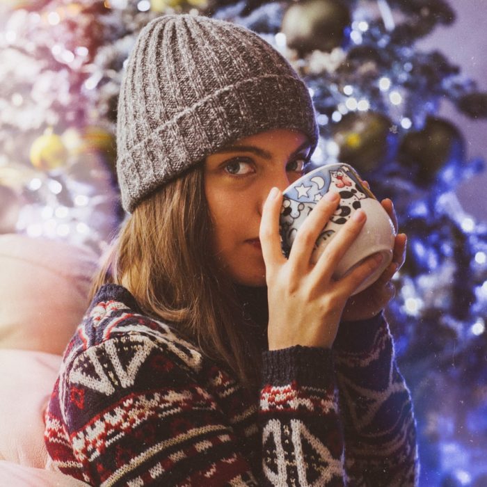 Chica con sueter navideño y gorro tejido para el frío, bebiendo cafpe en una taza de gatos frente al árbol de navidad con luces azules 