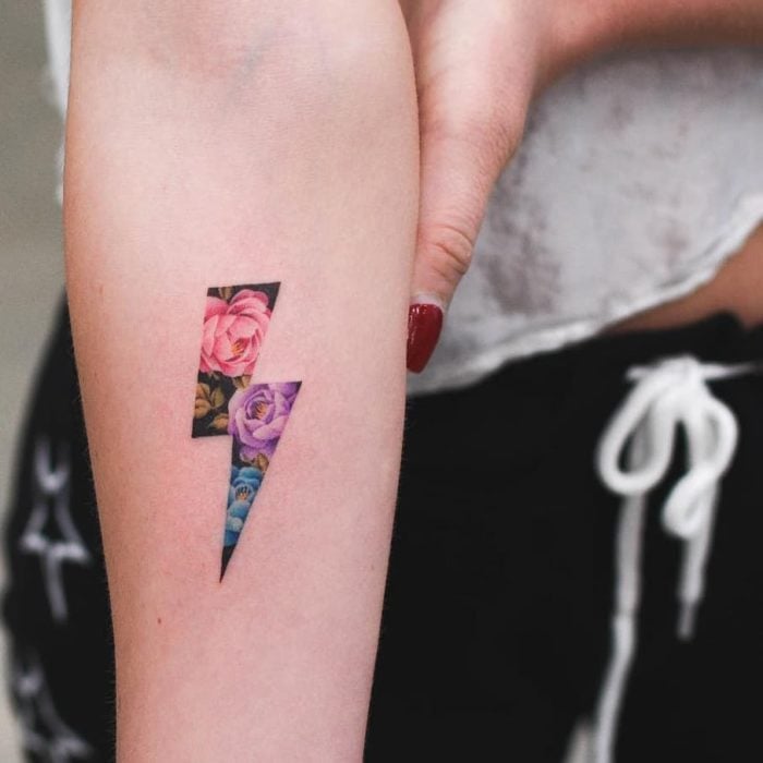 Tatuaje pequeño antebrazo en forma de rayo relleno de lores de colores y fondo negro