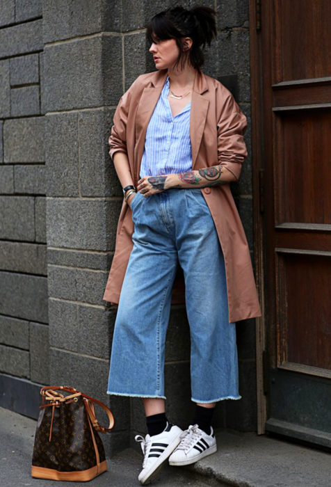 Looks con baggy jeans; chica en el pórtico de un edificio, con saco largo, tenis blancos y pantalón holgado, tatuajes en los brazos, peinado de media cola de cabello corto