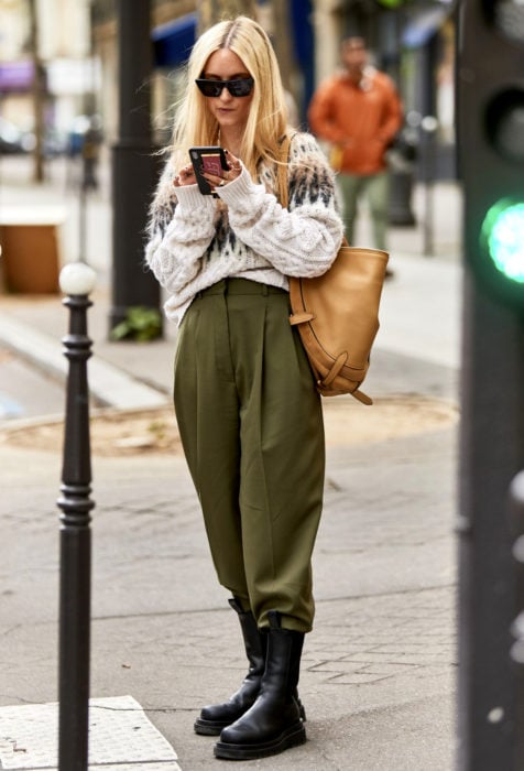 Looks con baggy jeans; mujer rubia mirando su celular en la calle, lentes oscuros, suéter tejido holgado, pantalón verde olivo oversized y botas negras