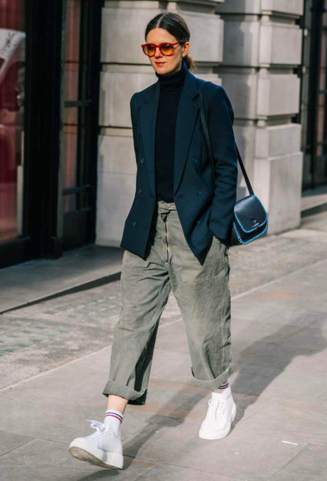 Looks con baggy jeans; mujer caminando en la calle con lentes rojos para el sol, sacp de vestid, pantalones oversized y tenis blancos