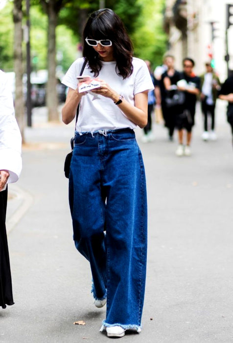 Looks con baggy jeans; mujer caminando en la calle mirando su celular, con blusa blanca básica y pantalones holgados