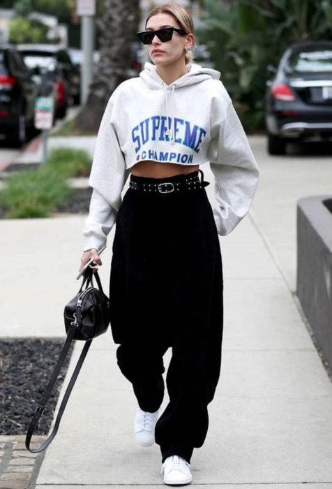 Looks con baggy jeans; Hailey Bieber caminando en la calle con hoodie crop top oversized, pantalones negros holgados con cinto de estoperoles y bolsa de mano