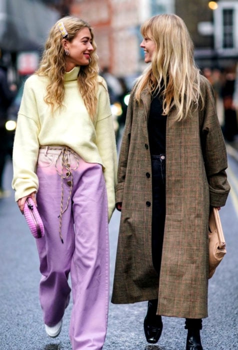 Looks con baggy jeans; amigas caminando en la calle y riendo, mujeres rubias con suéter oversized amarillo, pantalón holgado color lavanda, saco hasta los tobillos