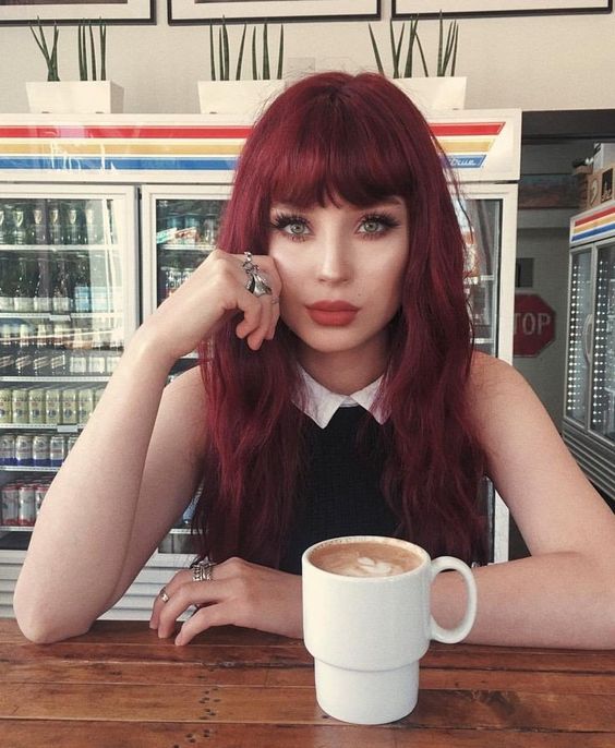 Chica sentada en uan cafetería, bebiendo café, mostrando su cabello largo tono cherry wine