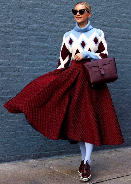 Outfits con medias de colores; mujer rucia con lentes de sol en forma de corazón, suéter oversized de rombos, falda midi rosa, medias azules y zapatos de charol color vino