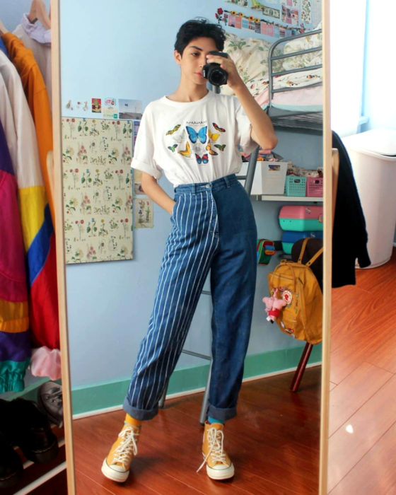 celestialyouth, joven recrea atuendos vintage de Sailor Moon; chico tomándose selfie frente al espejo con cámara profesional, playera blanca con estampado de mariposas, pantalón con una pierna rayada y la otra lisa, converse anaranjados