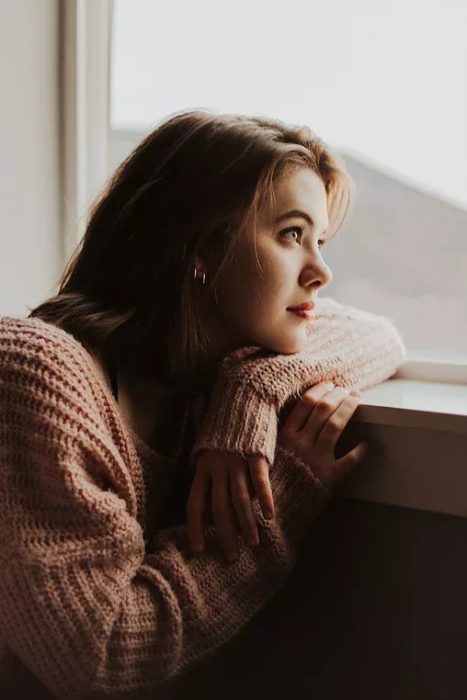 Mujer recargada sobre ventana mirando hacía afuera, vistiendo un suéter rosado