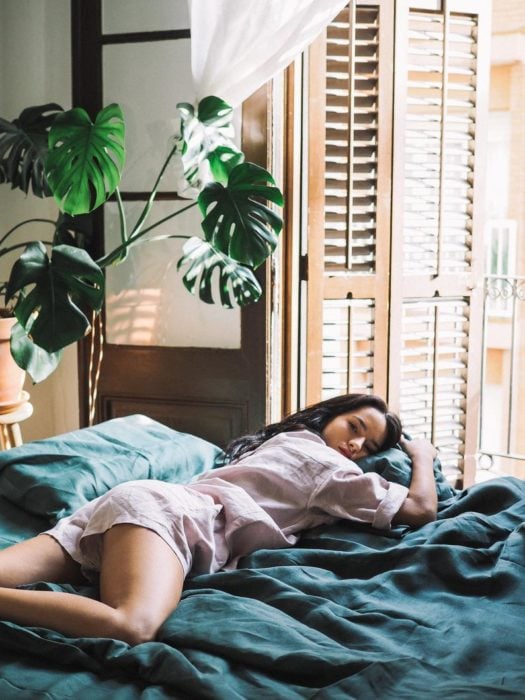 Chica recostada en la cama, usando pijama al lado de una ventana abierta