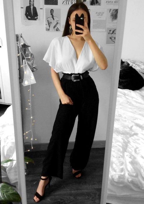 Atuendos formales y juveniles para oficina; mujer tomándose selfie frente al espejo, blusa blanca holgada, pantalón de vestir negro y cinto estilo vaquero