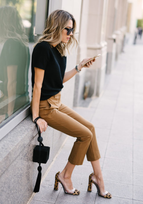 Atuendos formales y juveniles para oficina; mujer sentada en la calle, con lentes de sol, blusa negra, pantalones caqui y zapatos mule con animal print de leopardo