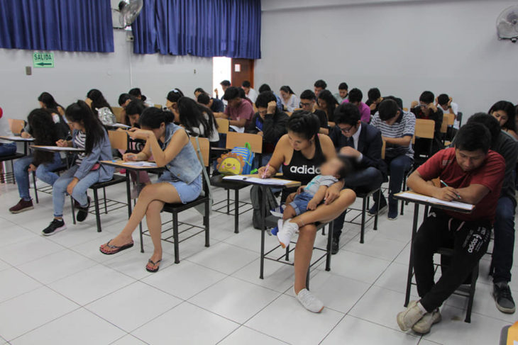 Chica sosteniendo en brazos a su bebé mientras contesta un examen en una aula de escuela 