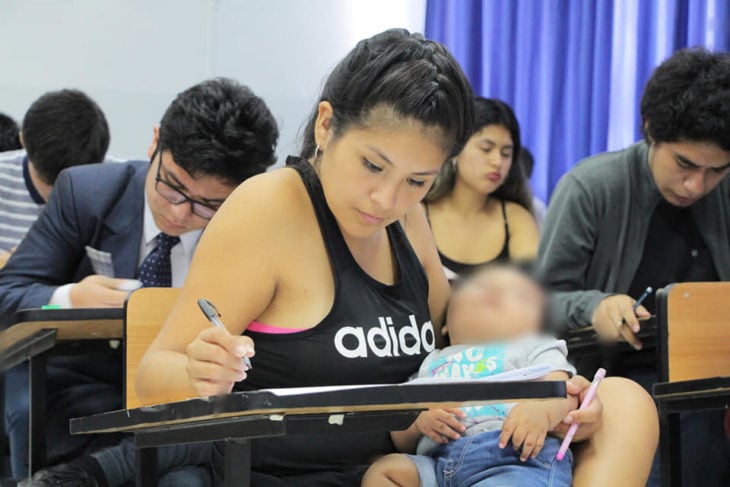 Chica sosteniendo en brazos a su bebé mientras contesta un examen en una aula de escuela 