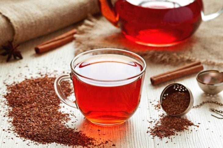 Té rooibos en taza de cristal adornado con canela, hojas trituradas de árbol de té e infusor de té 