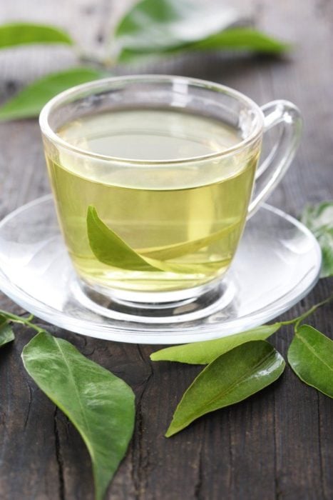 Té verde en taza de cristal sobre mes de madera oscura con hojas de de té adordando