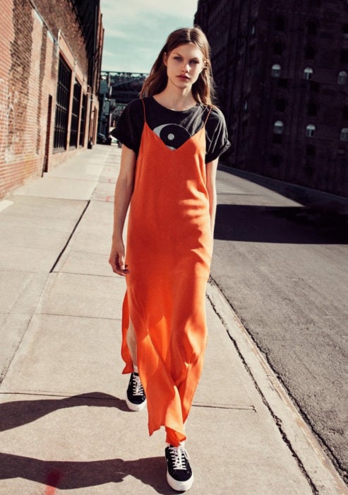 Looks con vestidos y blusas básicas; mujer caminando en la calle con maxivestido anaranjado, blusa con estampado de yin y yang, tenis negros y cabello largo y despeinado