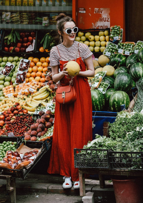 Looks con vestidos y blusas básicas; mujer en frutería en medio de frutas, con peinado de chongo alto, lentes de sol blancos, maxivestido rojo, blusa rayada, tenis