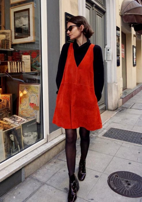 Looks con vestidos y blusas básicas; mujer en la calle viendo escaparates de tiendas, cabello corto y castaño, lentes de sol, vestido de gamuza rojo, con suéter de cuello alto y medias negras