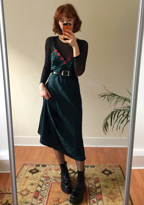 Looks con vestidos y blusas básicas; mujer tomándose selfie frente al espejo, vestido de satín verde con flores, blusa negra transparente, botas Dr. Martens, cabello rojo con corte de honguito y fleco