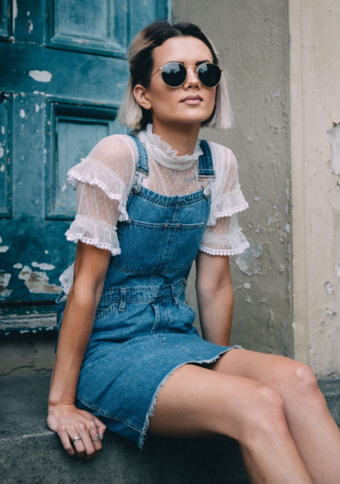 Looks con vestidos y blusas básicas; mujer sentada en escaleras de pórtico, cabello corto color blanco con raíces negras, lentes de sol, vestido overol, blusa de encaje y holanes blancos