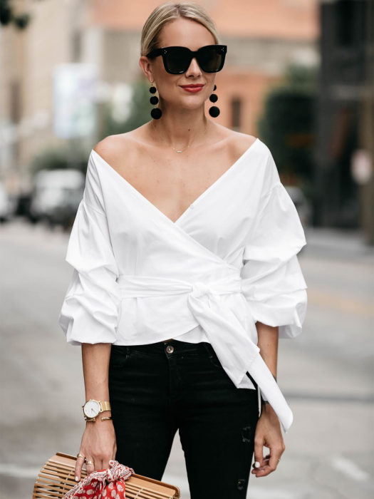 Wrap tops o blusas cache coeur blanca sin hombros; mujer rubia con peinado de coleta, lentes cuadrados grandes de sol, aretes negros colgantes y bolsa de madera