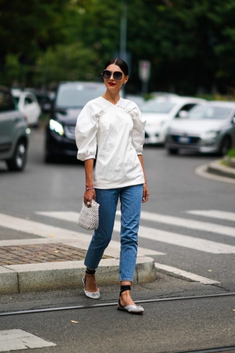 Mujer cruza la calle con blusa blanca y jeans oscuros mientras usa bailarinas blancas